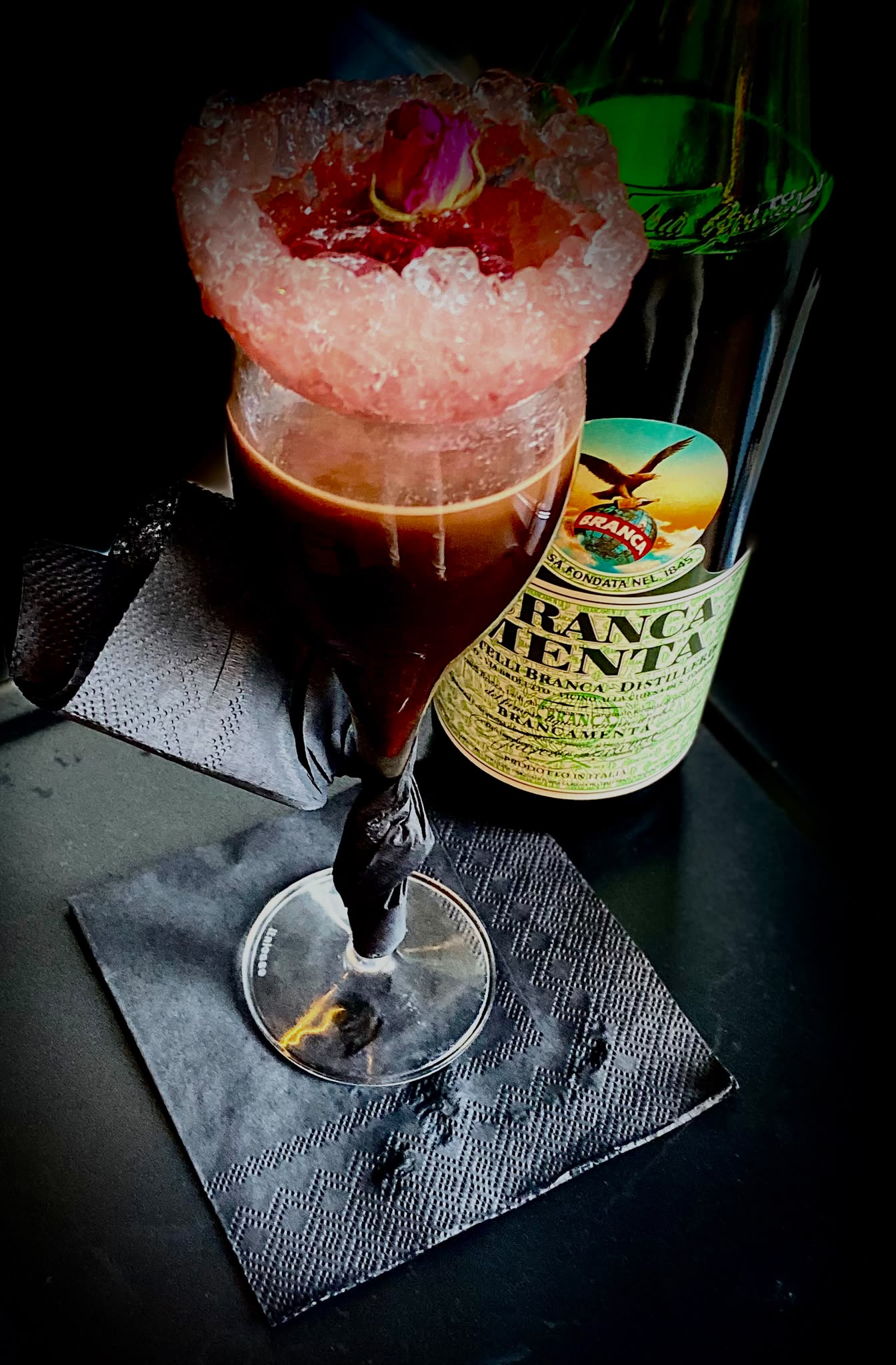Cocktail intens pe bază de cafea, rom, bitter aromat cu mentă, servit într-un pahar cu margine de gheață, zmeură confiată, floare comestibilă.